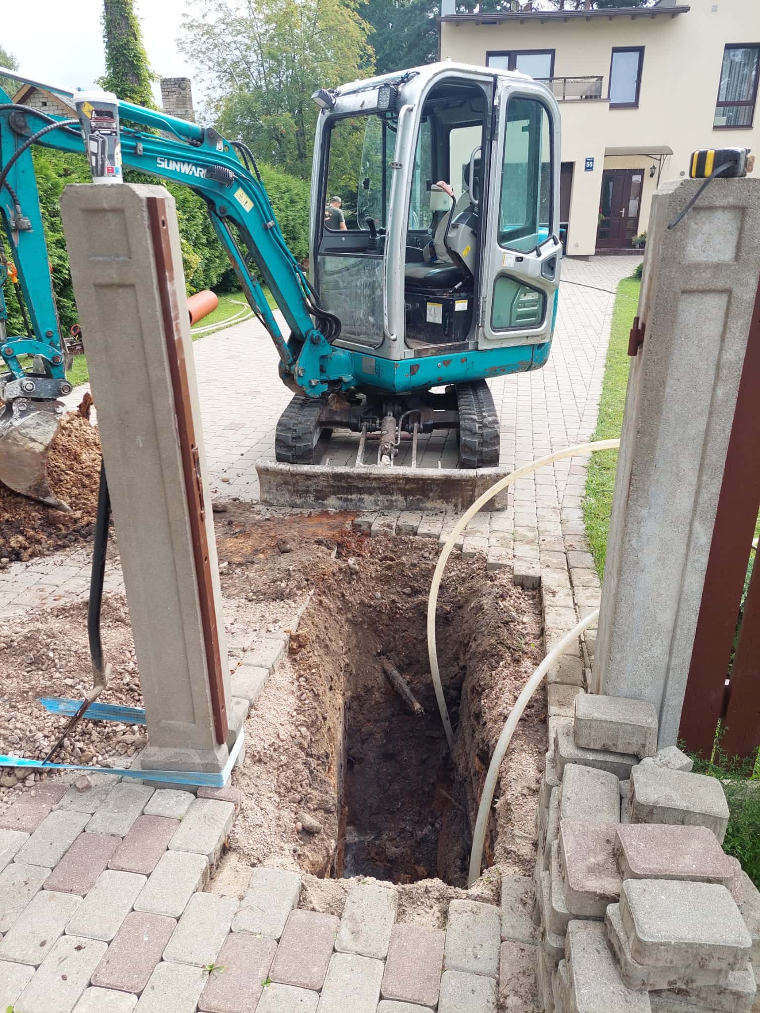 Mini ekskavators rok inženiertīklu tranšeju bruģētā privātmājas pagalmā